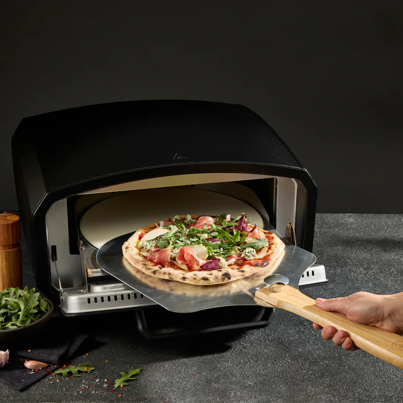 Person removing a pizza from a portable pizza oven on a dark surface.