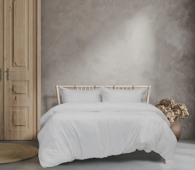 Neatly made bed with light-colored bedding in a room with wooden flooring and a textured wall.