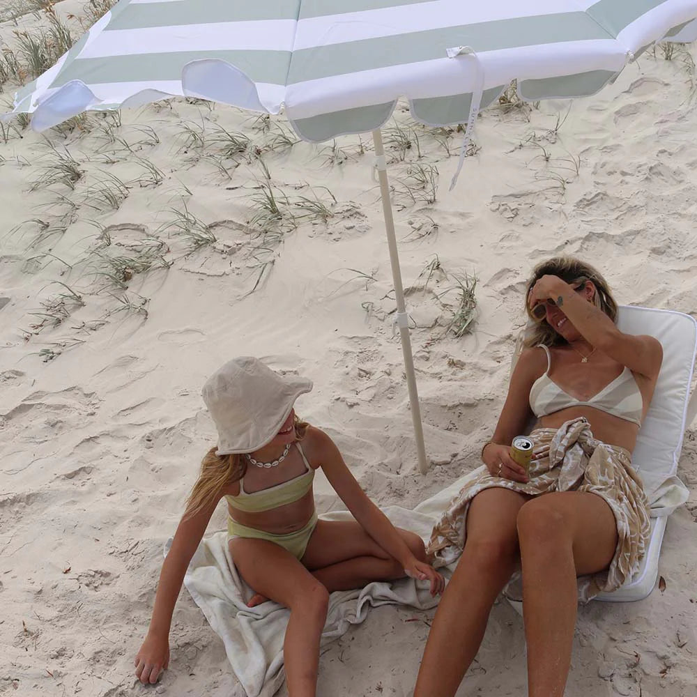 Two women sitting on a beach under a striped umbrella.