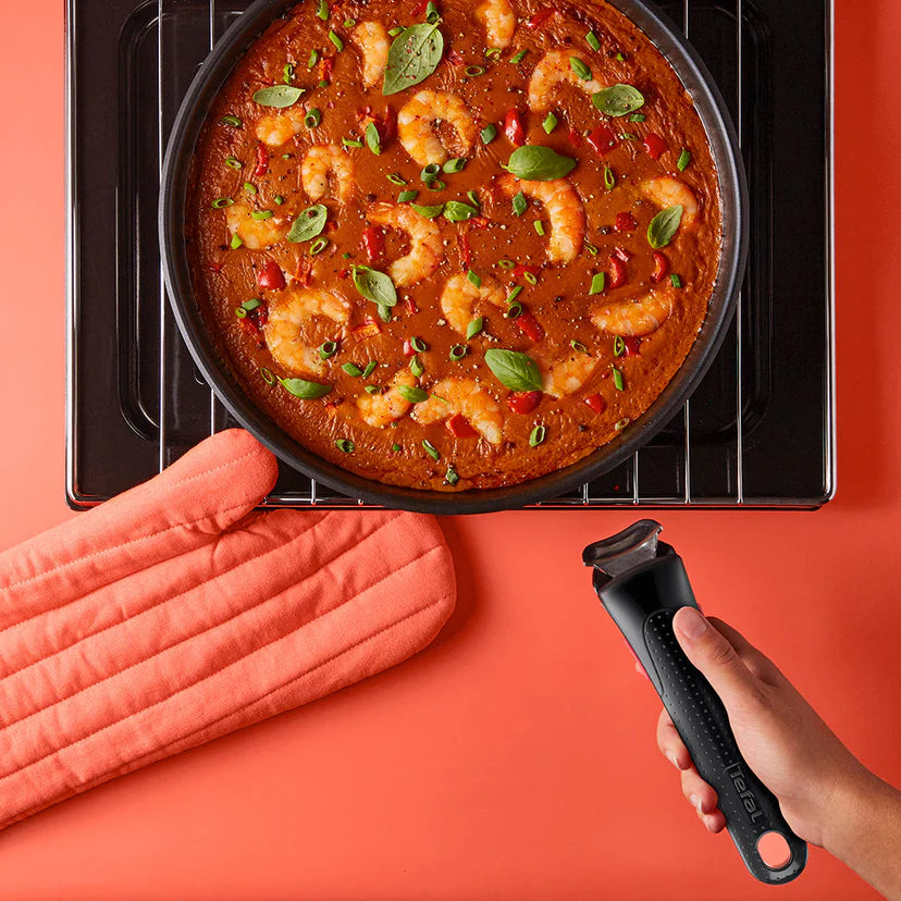 Frying pan with shrimp dish on a stove, person holding a kitchen tool, pink gloves on an orange background